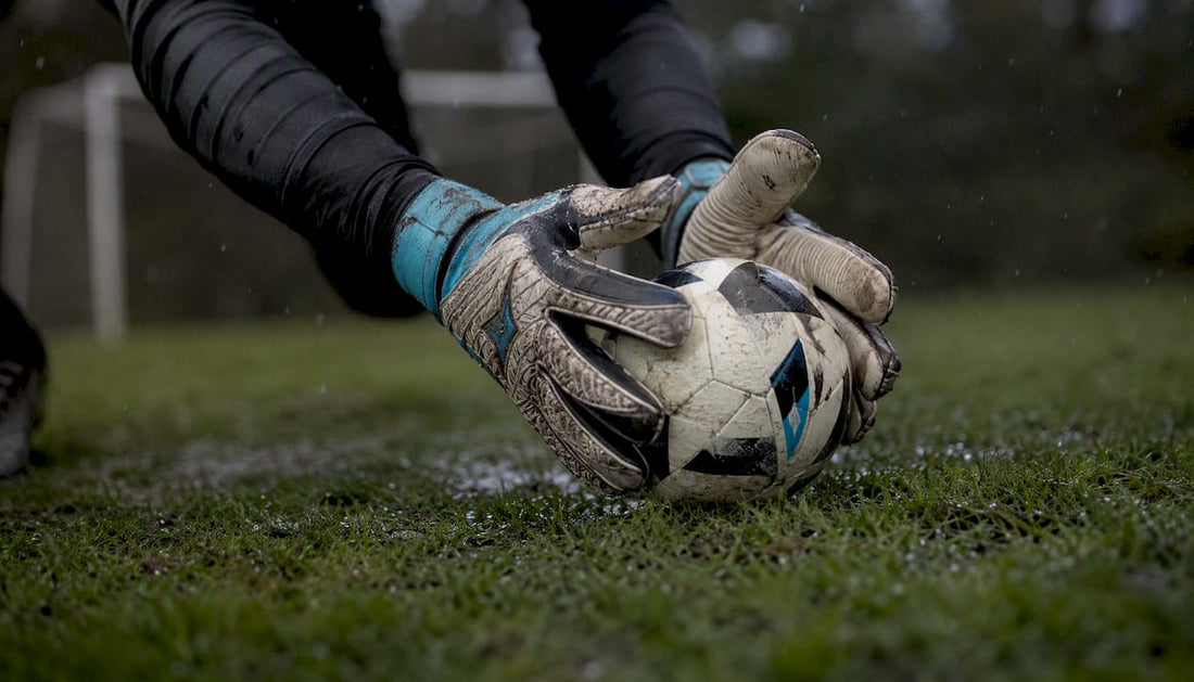 Mit Handschuhen greift der Torwart nach dem nassen Fußball auf dem vom Regen durchweichten Rasen.