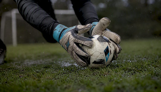 Mit Handschuhen greift der Torwart nach dem nassen Fußball auf dem vom Regen durchweichten Rasen.